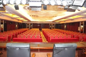 Un salón de conferencias con filas de sillas, un escenario con un atril y un techo con luminarias.
