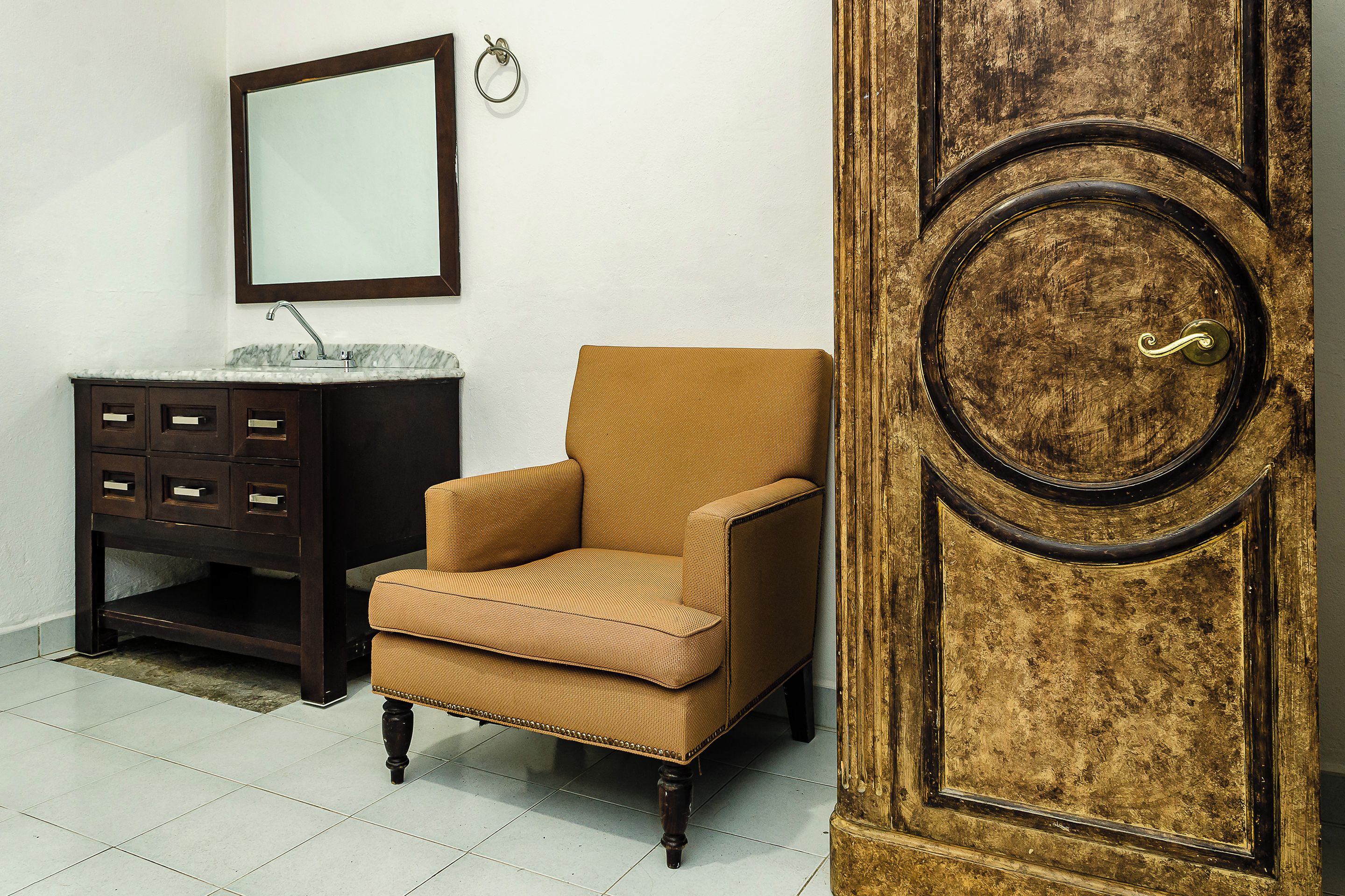 Una habitación con un sillón marrón, un aparador de madera oscura con cajones, un lavabo de mármol y una puerta de madera grande con manija de latón.