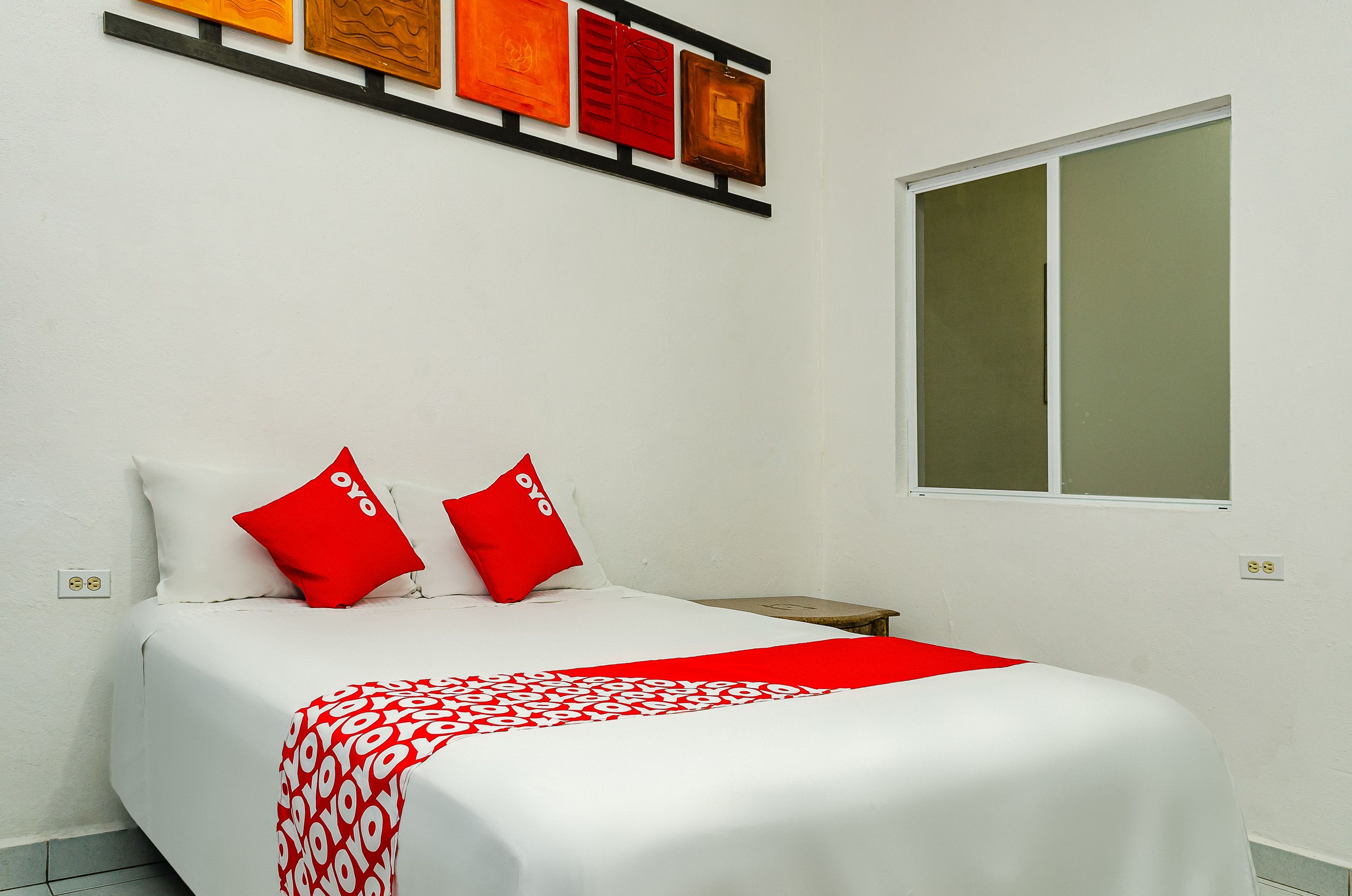 A neatly made bed with red and white bedding, a window with blinds, and a wall-mounted artwork above the bed.