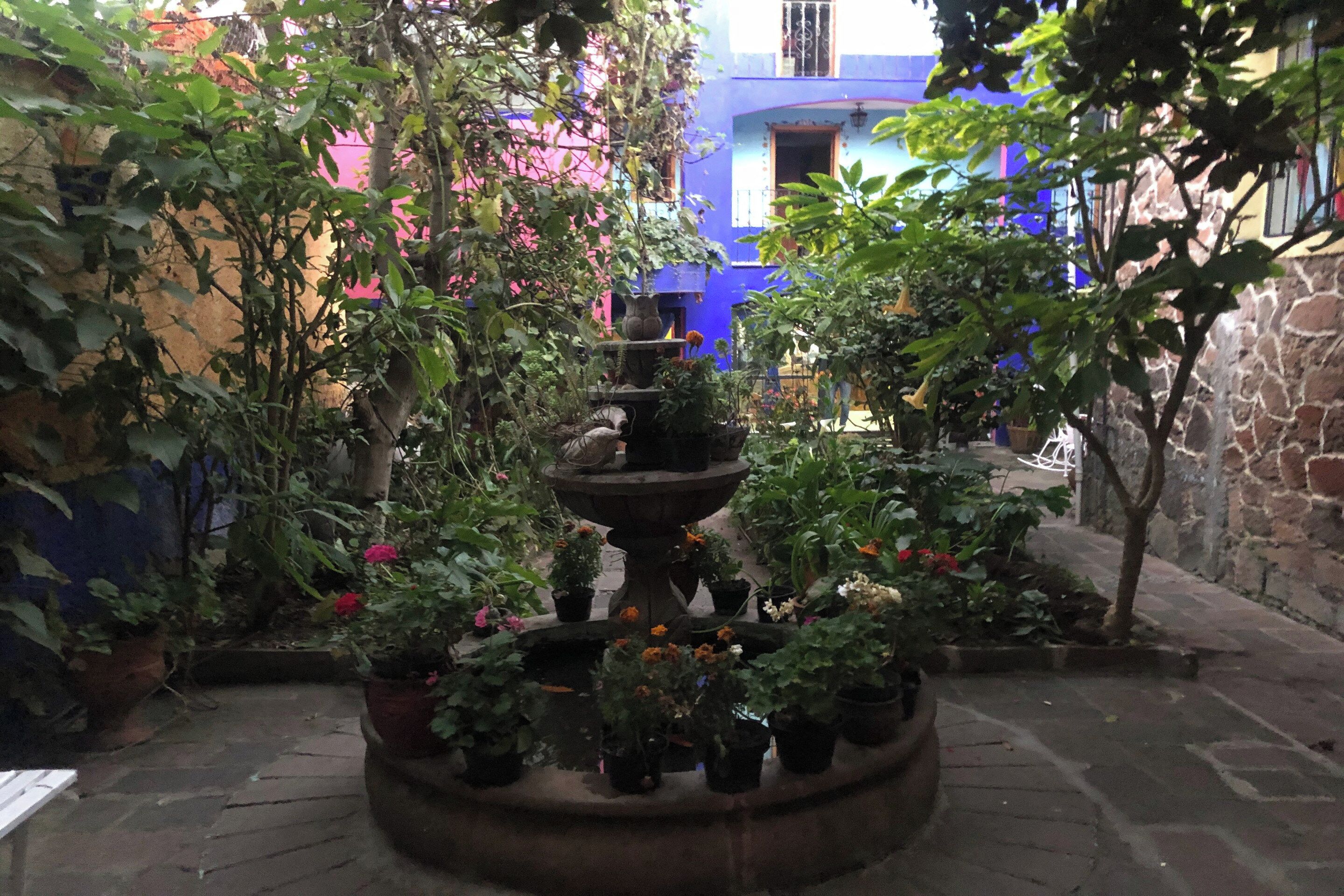 A courtyard with a central fountain surrounded by potted plants and a stone wall.
