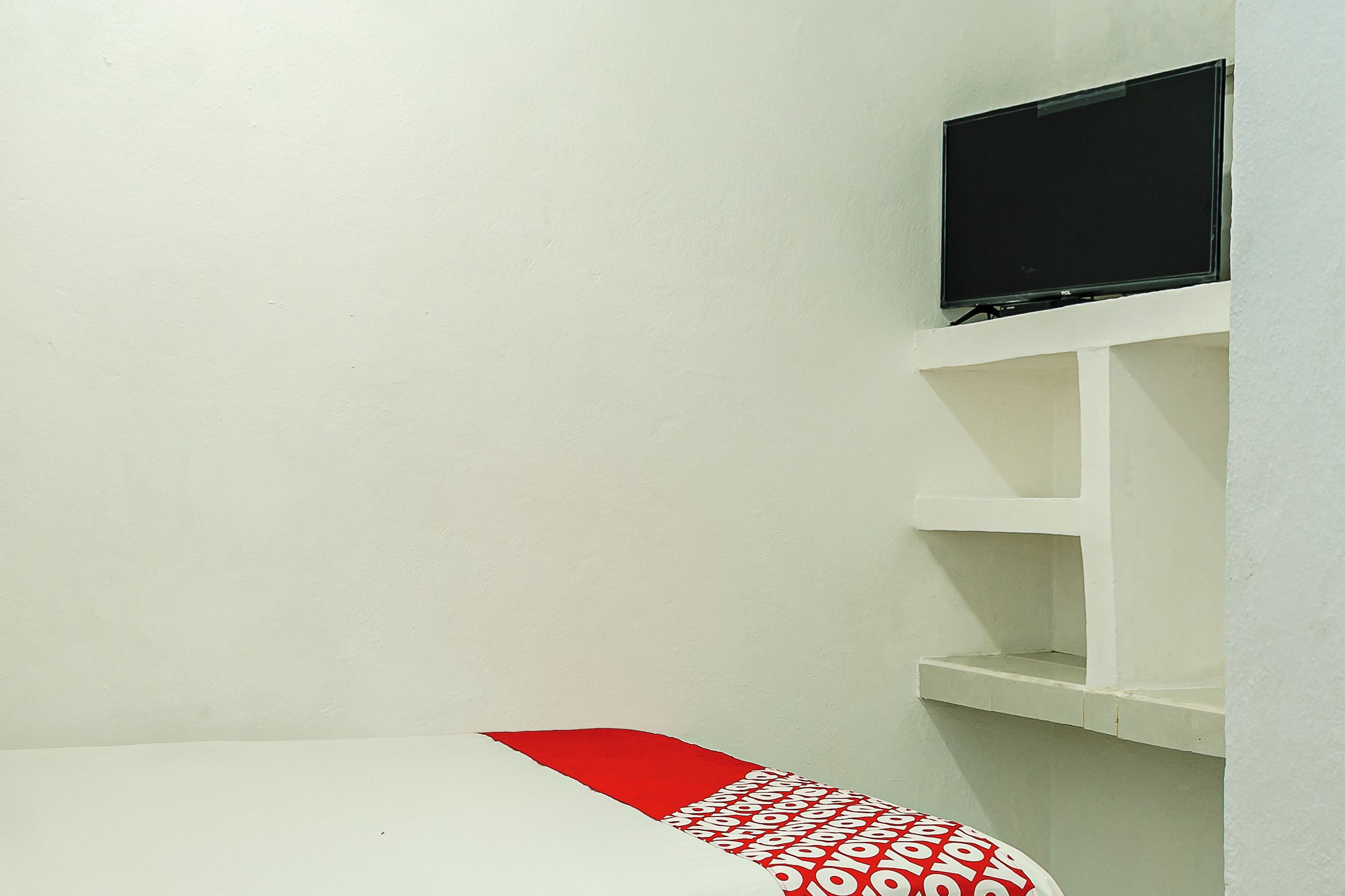 Um quarto pequeno e branco com uma cama coberta por um edredom vermelho e branco, uma televisão presa a uma estante branca acima da cama e uma parede branca.