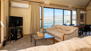 A hotel room with a large window, a flat-screen TV, a sofa, a dining table, and a small kitchenette.