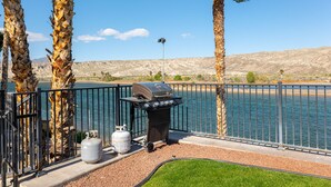 A lakeside barbecue setup with a grill and propane tanks, surrounded by palm trees and a metal fence.