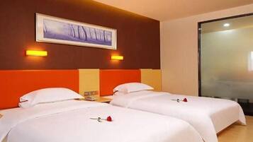 A hotel room with two beds, each with white bedding and a red rose on the bed. There is a painting on the wall above the beds and a glass partition on the right side.