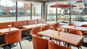 A modern restaurant with orange seating, large windows, and a view of buildings. - EVEN Hotel Long Island City - New York by IHG (Long Island City)