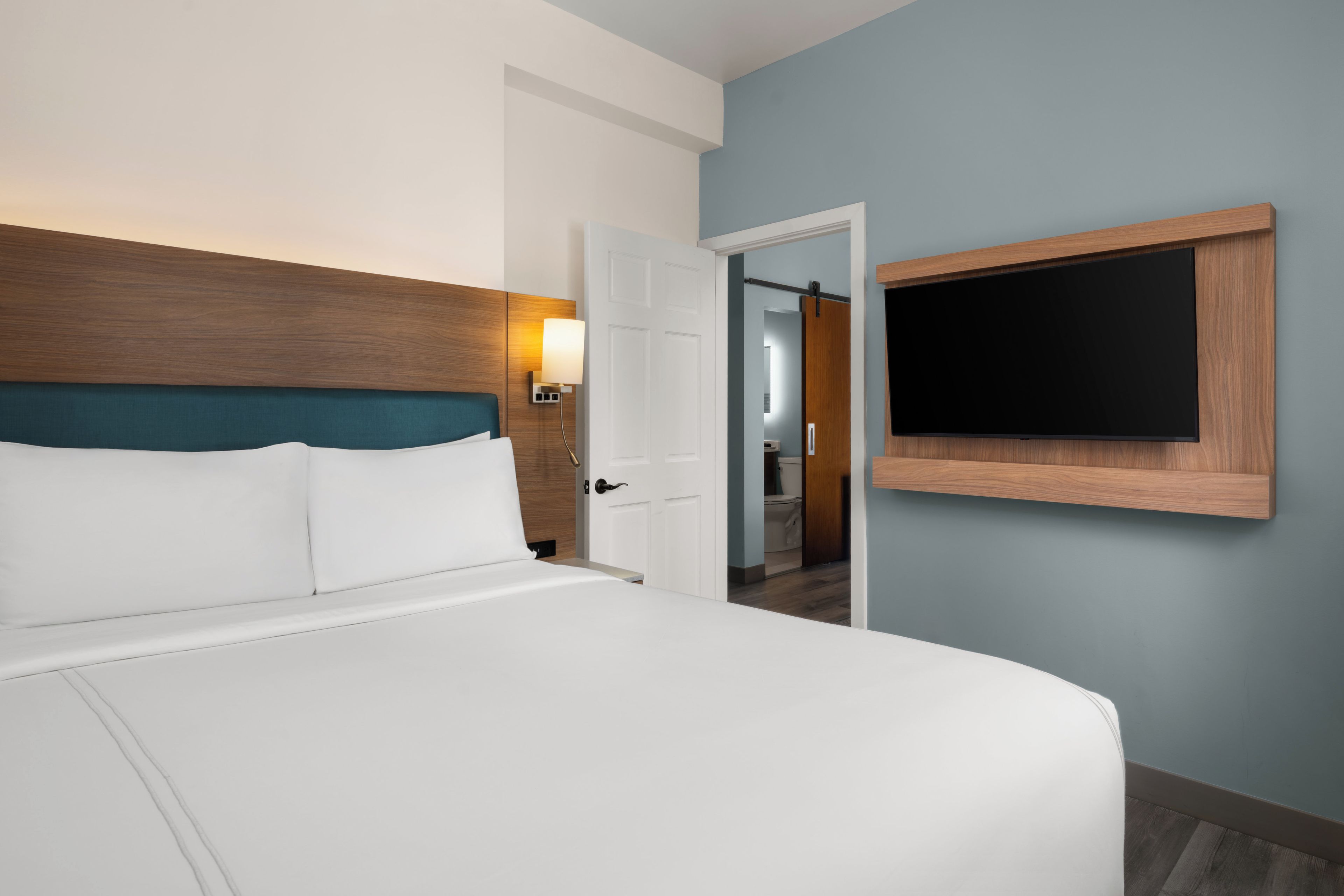 Ein Hotelzimmer mit Bett, einem an der Wand befestigten Fernseher und einer Tür zu einem Badezimmer.