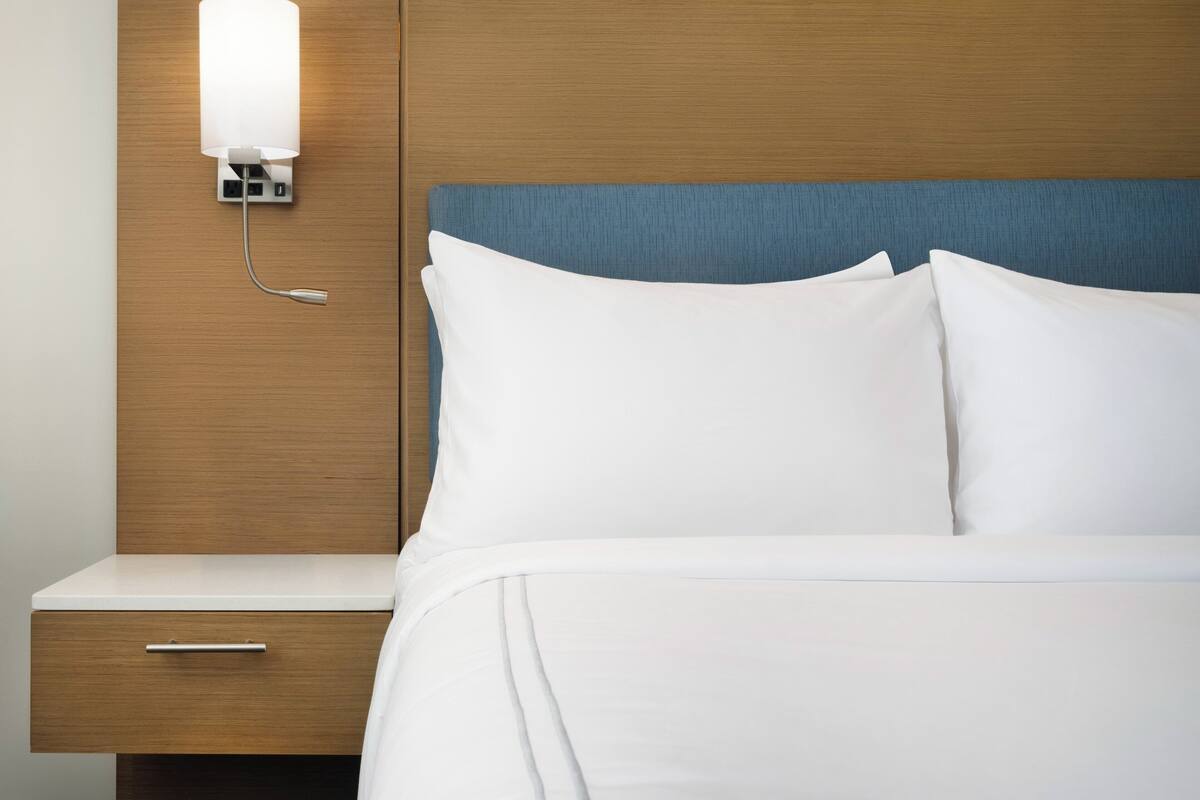 Une chambre d’hôtel avec une tête de lit en bois, une lampe de chevet, un lit blanc et une table de chevet en bois dotée d’un tiroir.