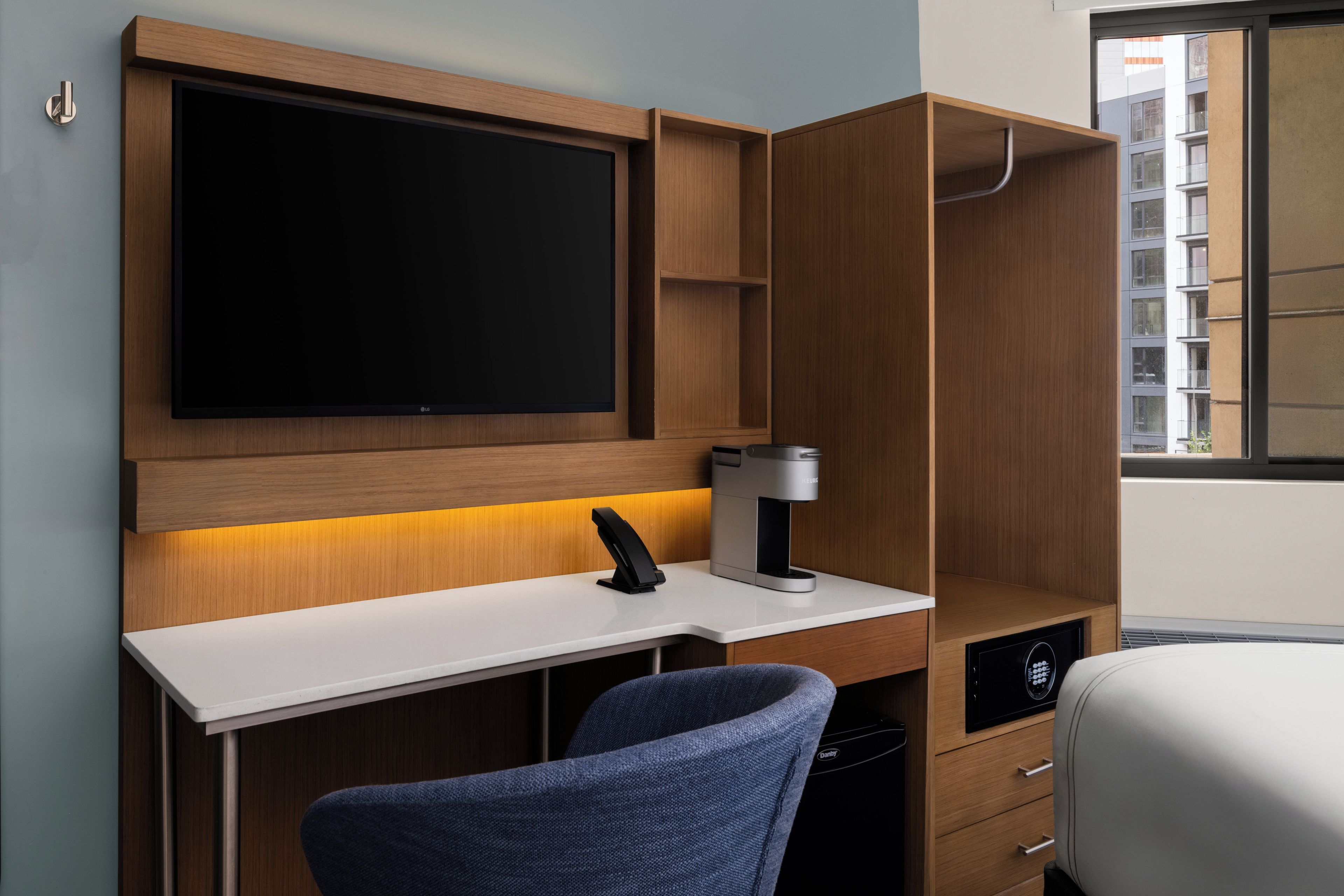 A modern hotel room with a flat-screen TV mounted on a wooden wall, a desk with a coffee machine, a chair, and a built-in dishwasher.