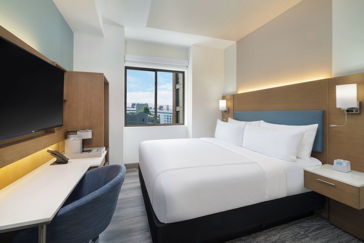 A hotel room with a bed, a desk, a chair, a television, and a window with a city view.