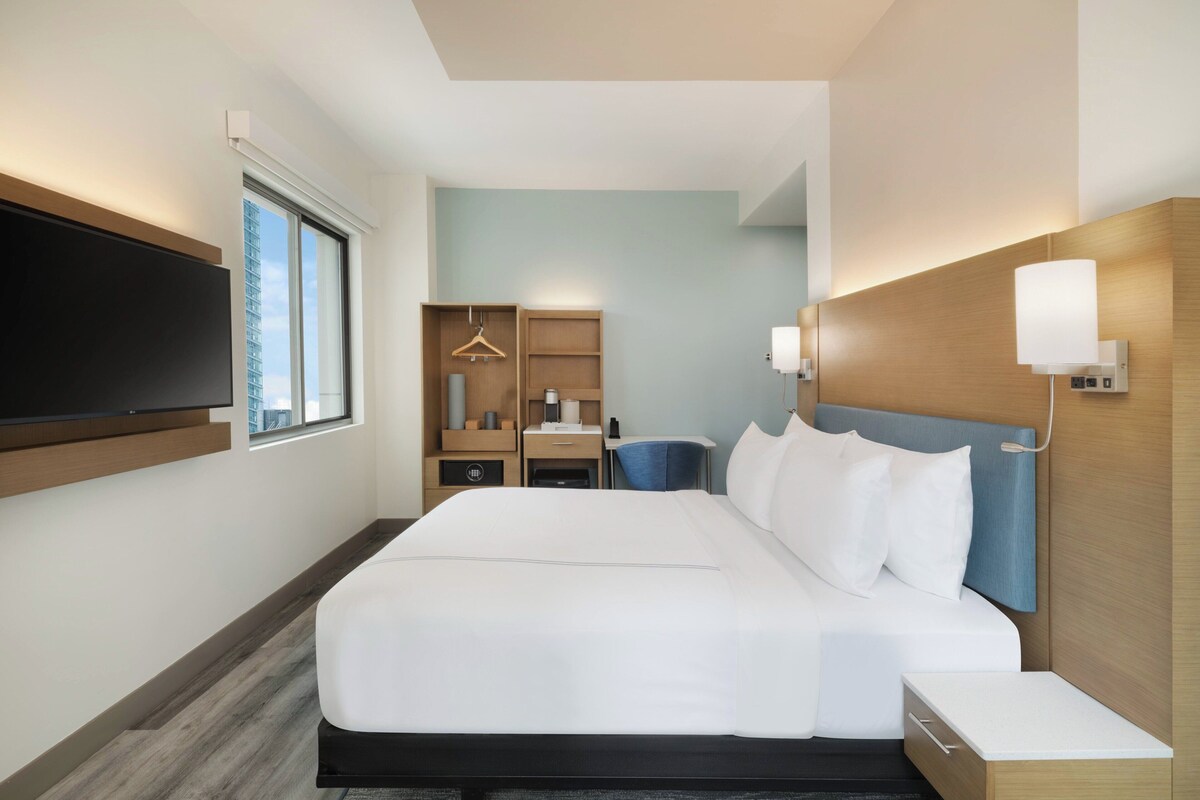A hotel room with a large bed, a desk, a chair, a TV, and a wardrobe.
