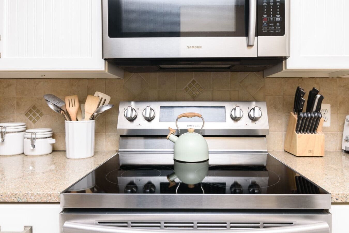 Una cocina con electrodomésticos de acero inoxidable, incluyendo un microondas, horno y estufa. Hay utensilios en un recipiente, una tetera sobre la estufa y un juego de cuchillos en la encimera.