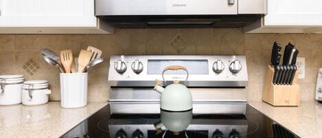 Una cocina con electrodomésticos de acero inoxidable, incluyendo un microondas, horno y estufa. Hay utensilios en un recipiente, una tetera sobre la estufa y un juego de cuchillos en la encimera.