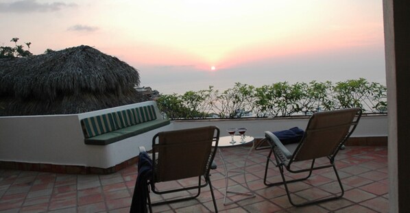 Terraço com duas cadeiras e uma mesa, com vista para um pôr do sol e uma construção de palha.