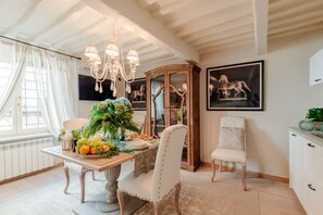 A dining area with a wooden table set for four, surrounded by upholstered chairs. A chandelier hangs above, and there are framed pictures on the wall.