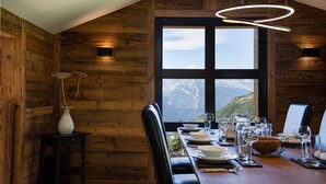 A dining area with a wooden interior, a large window offering a mountain view, and a modern pendant light. - Chalet Lupin - Four Bedroom Chalet, Sleeps 11 (Riddes)