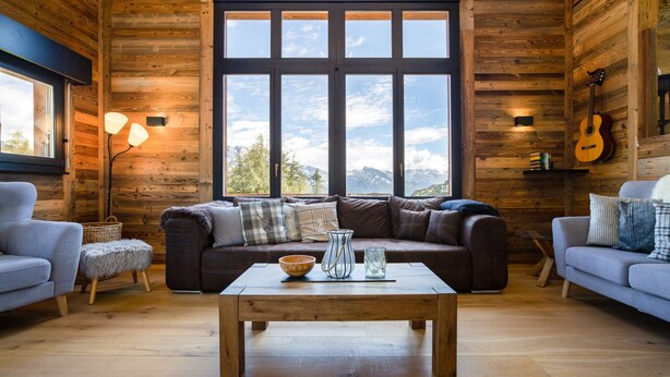 A cozy log cabin interior with a large window, a wooden coffee table, and two sofas. - Chalet Lupin - Four Bedroom Chalet, Sleeps 11 (Riddes)