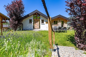A chalet-style building with a sloped roof, a wooden deck, and a stone pathway leading to the entrance. - Chalet Lupin - Four Bedroom Chalet, Sleeps 11 (Riddes)