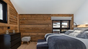 A bedroom with a wooden wall, a bed with grey bedding, a small stool, and a dresser. - Chalet Lupin - Four Bedroom Chalet, Sleeps 11 (Riddes)
