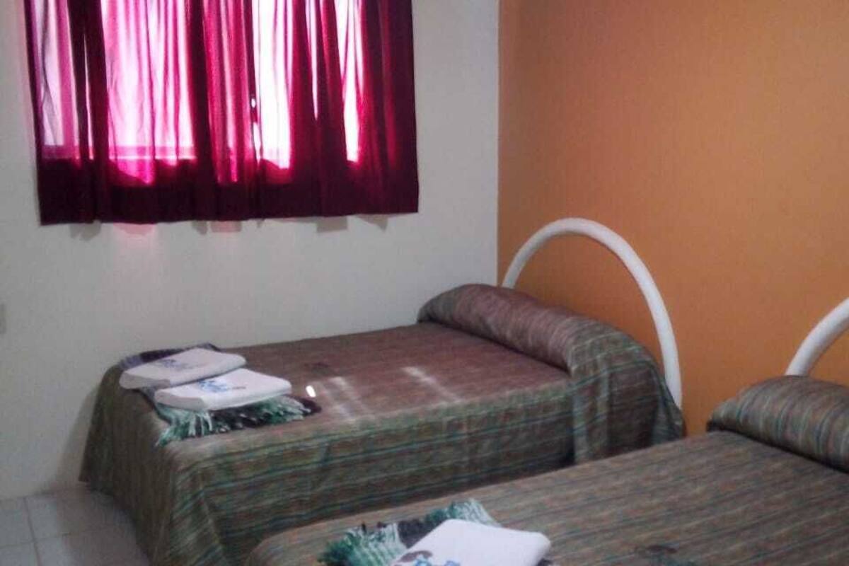 Une chambre avec deux lits simples, chacun doté d’une tête de lit et d’un oreiller. Il y a une fenêtre avec des rideaux rouges, ainsi qu’une petite table et une chaise.
