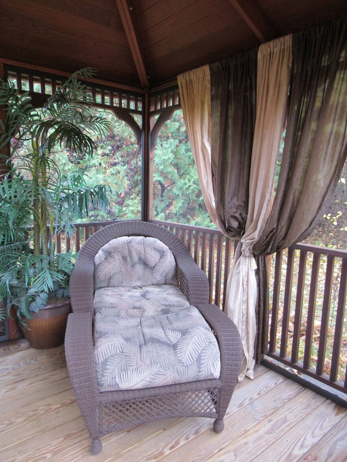 A wicker chair with a leaf-patterned cushion on a wooden deck, surrounded by potted plants and a wooden pergola with curtains.