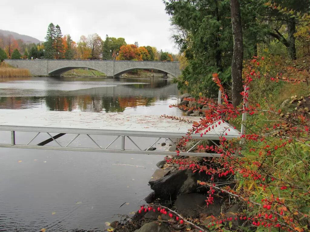 Un puente de piedra sobre un río tranquilo, con follaje otoñal y una barandilla de metal a lo largo de la orilla.