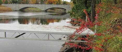 Un puente de piedra sobre un río tranquilo, con follaje otoñal y una barandilla de metal a lo largo de la orilla.