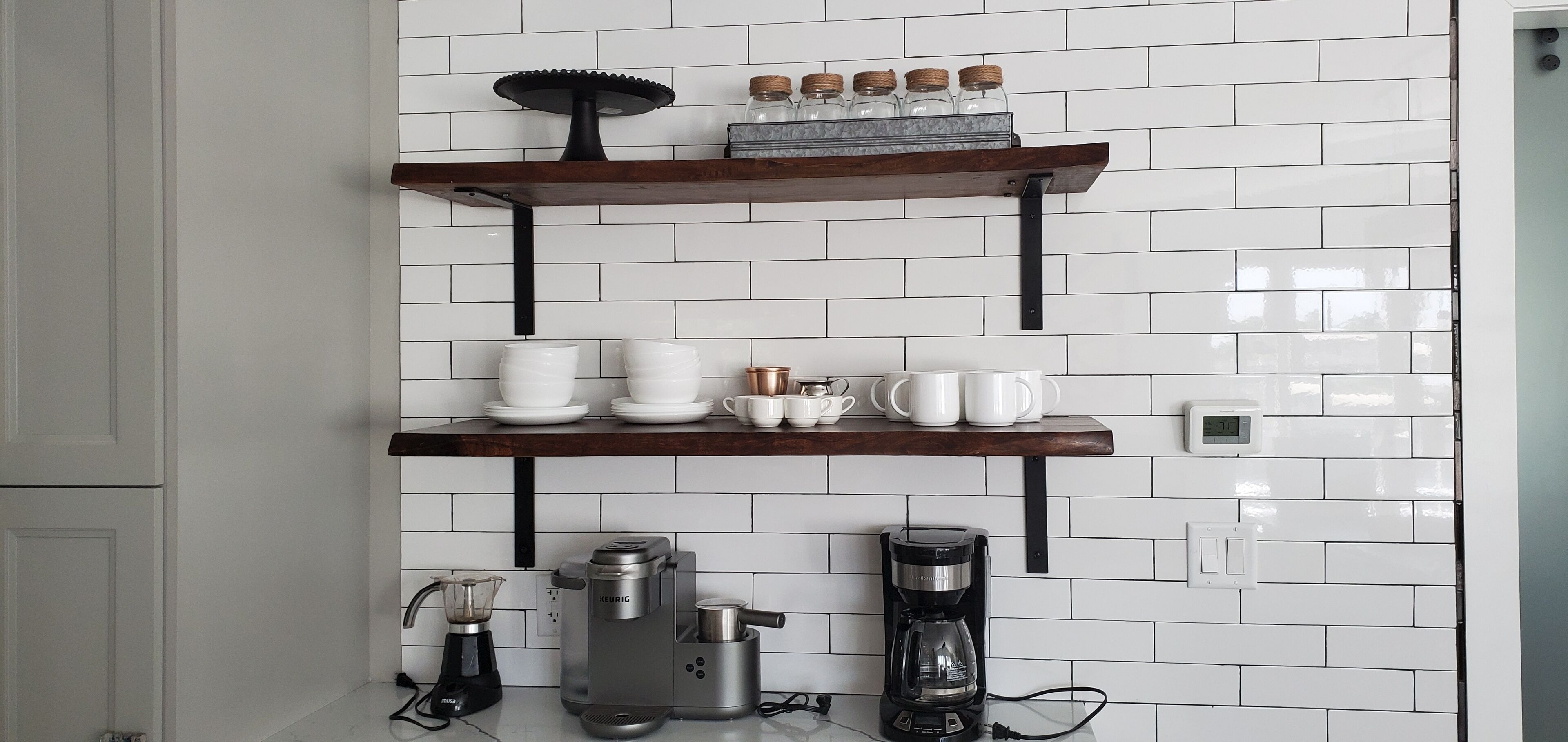A kitchen with white subway tiles, two wooden shelves, and coffee makers.