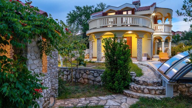 Una casa de dos pisos con un balcón curvo, paredes de piedra y un camino de piedra que lleva a la entrada.