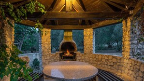 Un horno de pizza de piedra al aire libre con fuego encendido, rodeado de muros de piedra y una estructura de techo de madera.