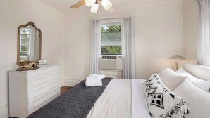 Un dormitorio con cama, cómoda, ventilador de techo, ventana con cortinas y vista a zonas verdes.
