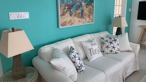 A teal-colored room with a white sofa, decorative pillows, wall-mounted lamps, and a framed painting of a sea turtle. - Beachfront Apartment in Paradise: Turtles Nest D (Dover)