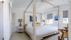 A four-poster canopy bed with a white bedspread and decorative pillows, a wooden nightstand with a lamp, and a view of the sea through a window with patterned curtains. - Beachfront Apartment in Paradise: Turtles Nest D (Dover)