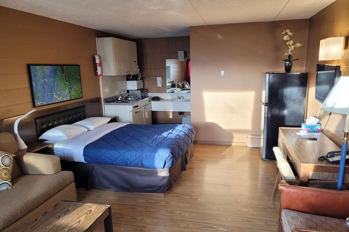 Habitación de hotel con cama, sofá, escritorio, televisor, refrigerador, microondas, cafetera y una pequeña cocina.