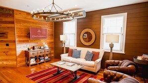 Un salon chaleureux avec un canapé blanc, un fauteuil en cuir, des murs en bois, un lustre et une enseigne portant l’inscription « Charlestonian ».