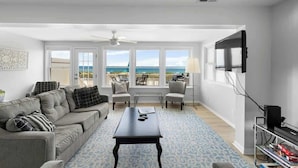 A living room with a sofa, chairs, a coffee table, a TV, and a balcony with a view. - Ocean Oasis by Carolina Getaway - Oceanfront Townhome w/ Private Heated Pool (Kure Beach)