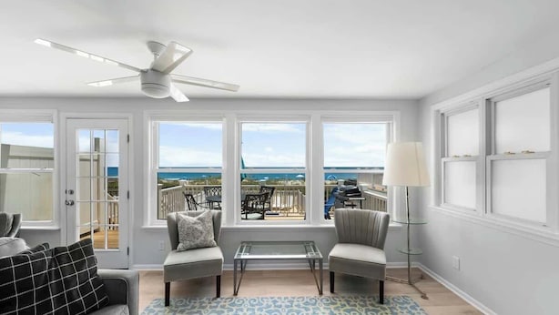A room with a ceiling fan, two sofas, a glass coffee table, a patterned rug, and large windows with a view of the beach. - Ocean Oasis by Carolina Getaway - Oceanfront Townhome w/ Private Heated Pool (Kure Beach)