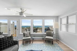 A room with a ceiling fan, two sofas, a glass coffee table, a patterned rug, and large windows with a view of the beach. - Ocean Oasis by Carolina Getaway - Oceanfront Townhome w/ Private Heated Pool (Kure Beach)