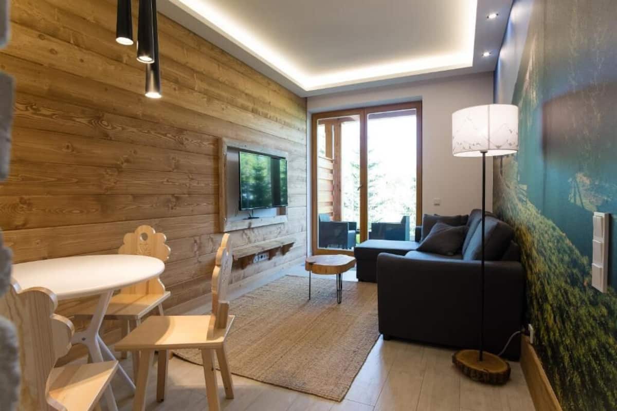 A cozy living room with a wooden wall, a flat-screen TV, a sofa, and a dining table with chairs.