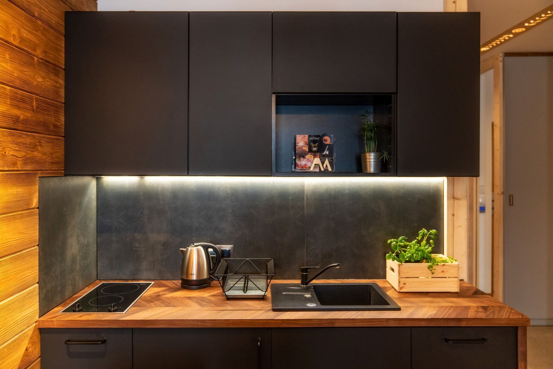 A modern kitchen with a wooden countertop, black cabinets, a built-in oven, and a small shelf with a plant and a magazine.