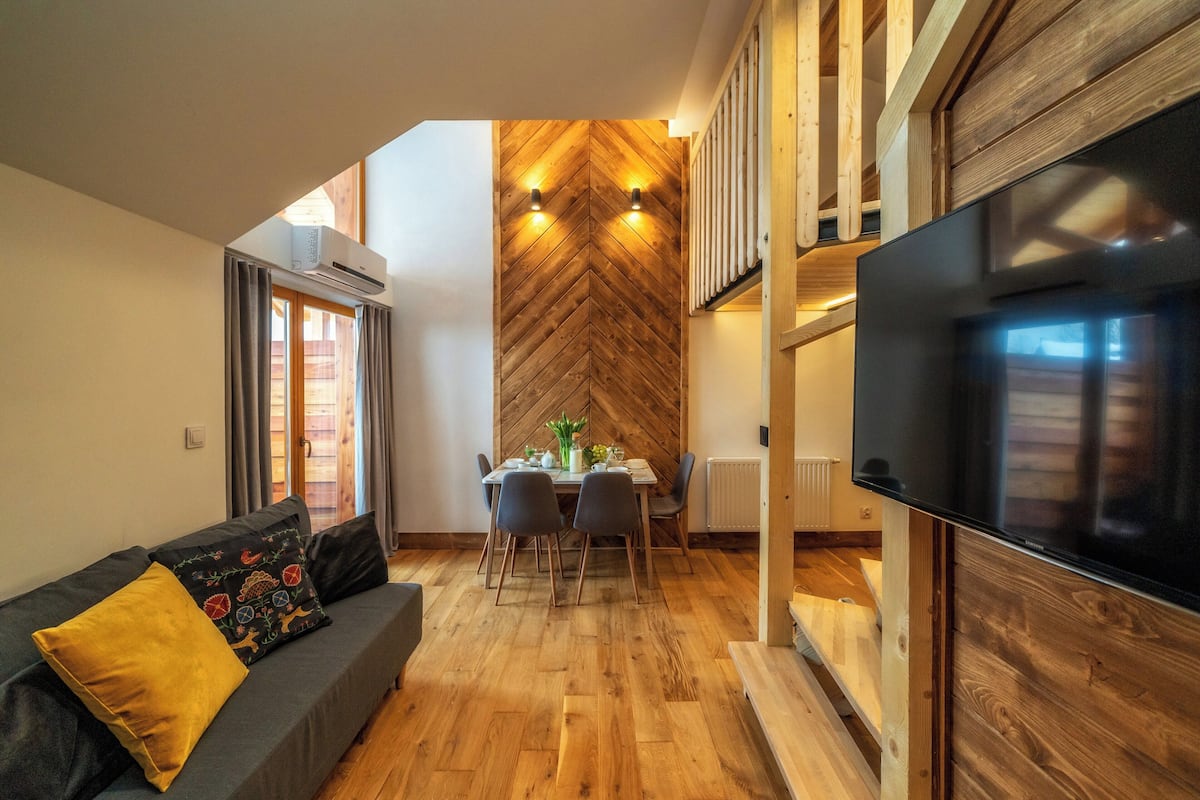 A modern living room with a herringbone wood wall, a dining area with a table and chairs, and a large flat-screen TV mounted on a wooden wall.
