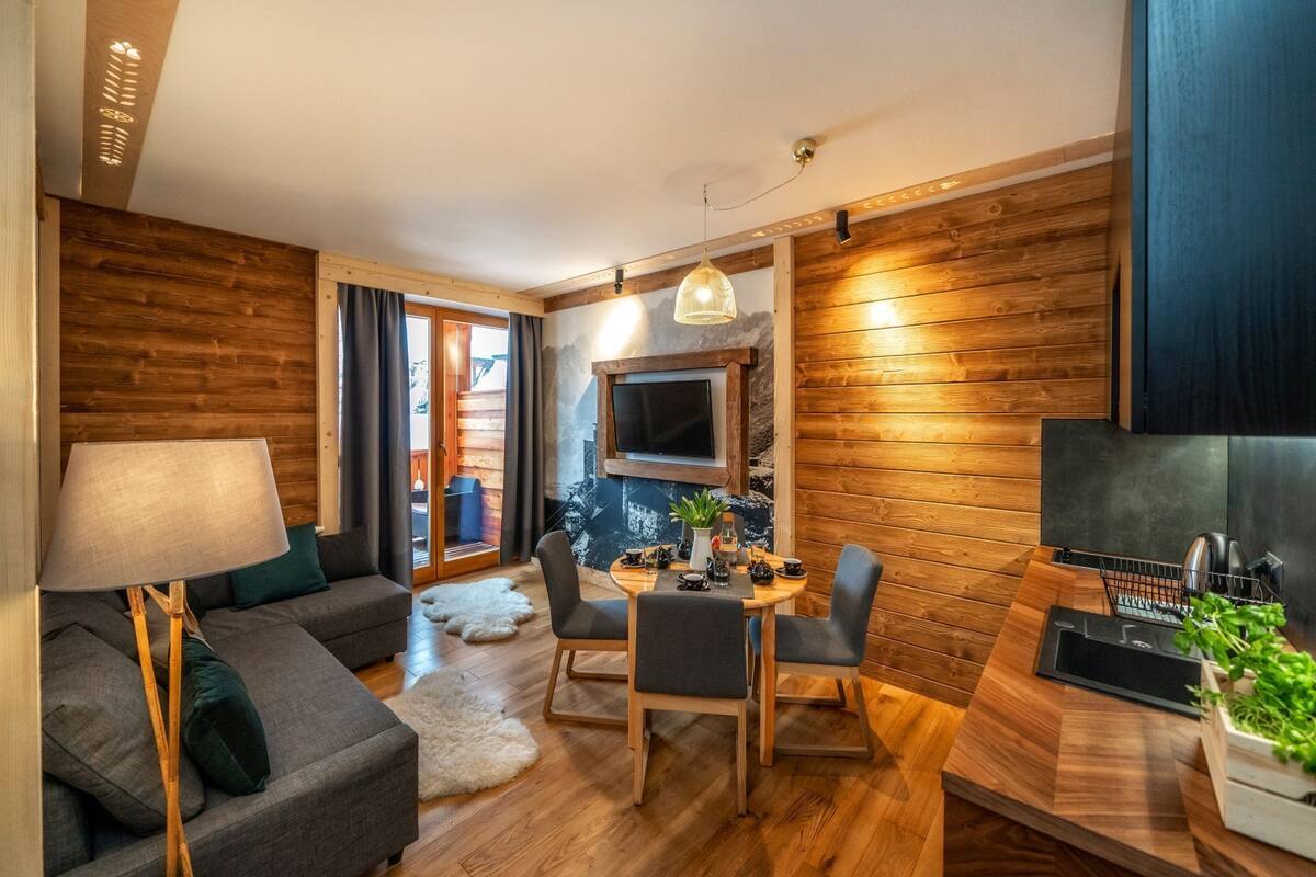 A cozy living room with a sofa, a dining table, and chairs. There's a TV mounted on the wall and a wooden floor.