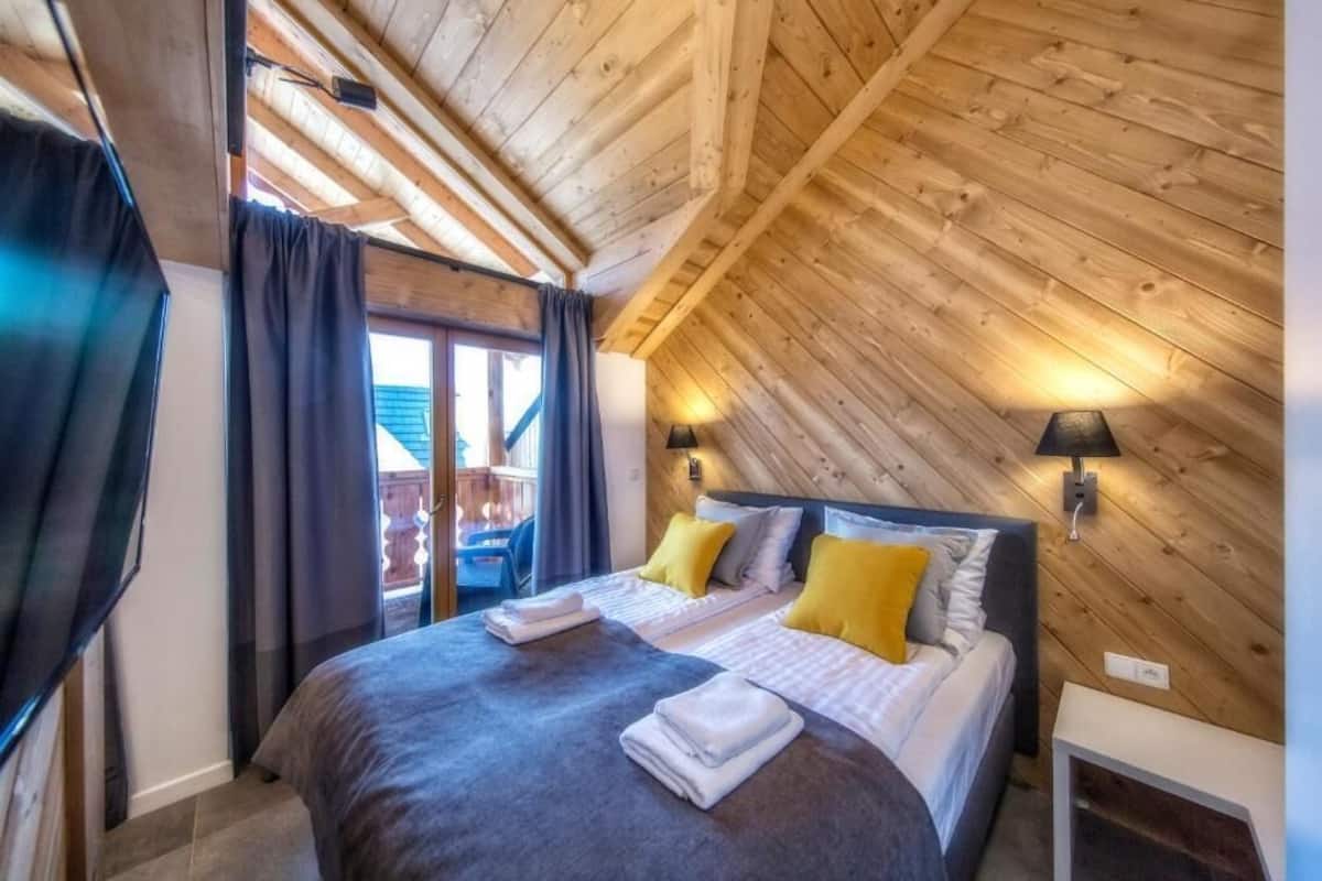 A cozy bedroom with a sloped ceiling, a bed with pillows, a TV, and a window with curtains.