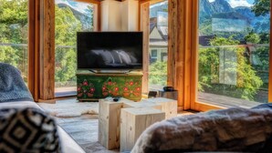 Interior de madera con un televisor sobre un soporte de madera, un manto de piel y vistas a montañas a través de amplios ventanales.