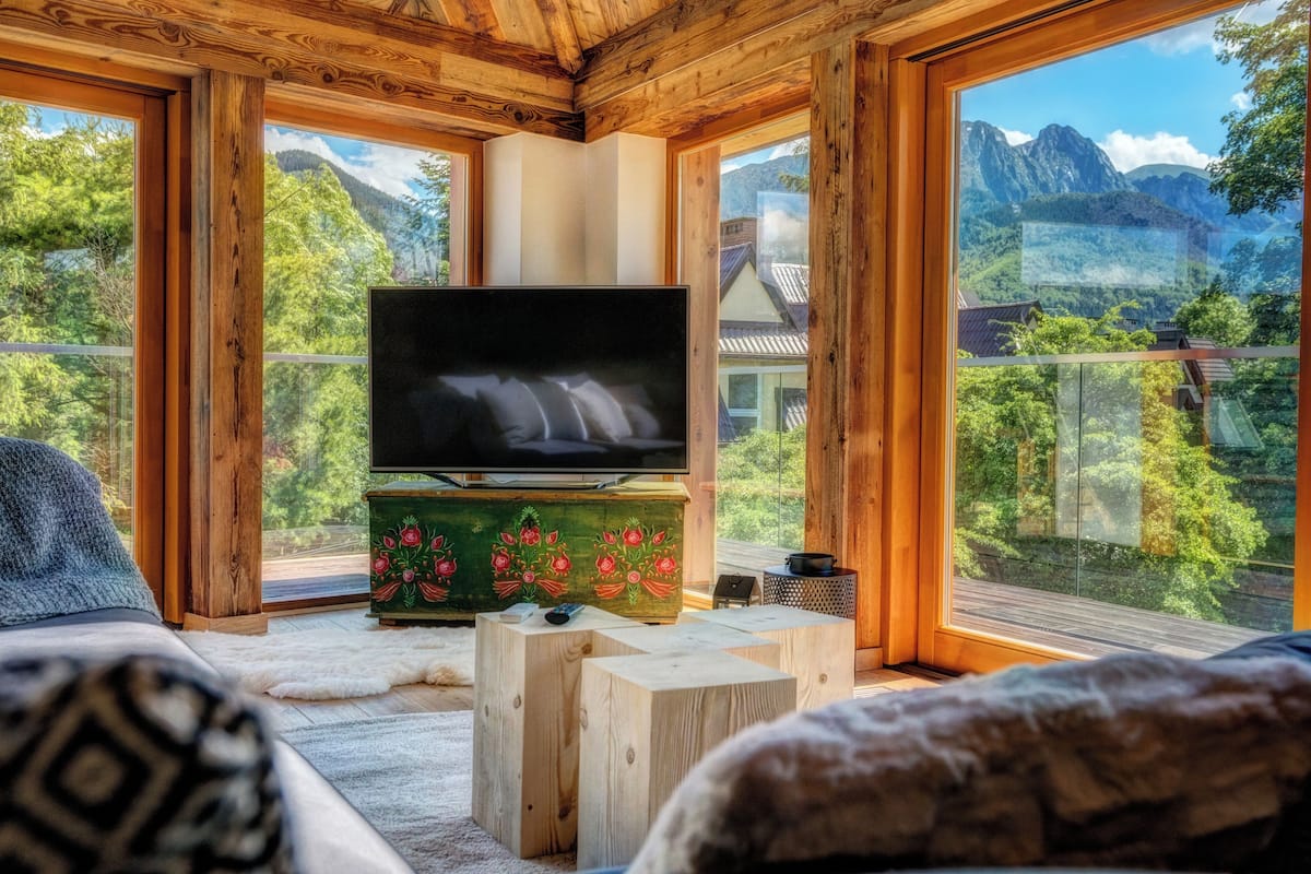 A wooden interior with a TV on a wooden stand, a fur rug, and a view of mountains through large windows.