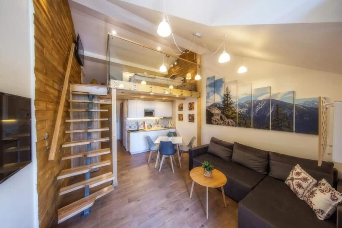 A modern apartment with a wooden staircase, a kitchen, a dining area, and a living room with a sofa and a small table.