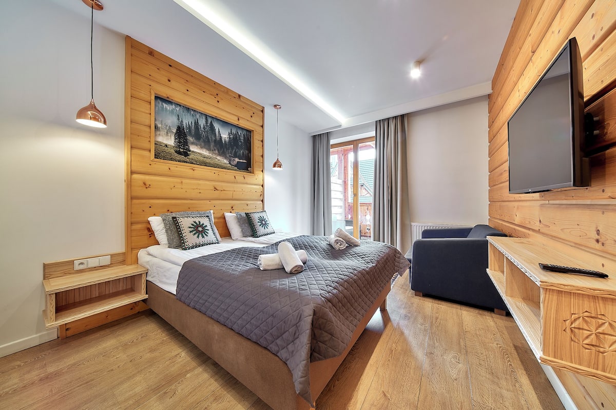 A modern bedroom with a wooden headboard, a large bed with a quilted blanket, a bedside table with a lamp, and a TV mounted on the wall.