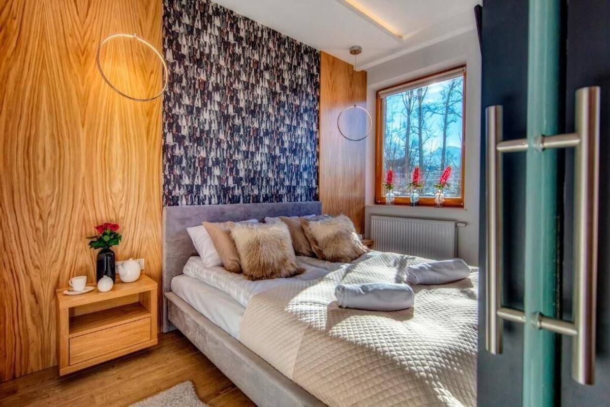 A bedroom with a wooden headboard, a patterned wall, a bedside table with a vase of flowers, and a window with a view of trees.