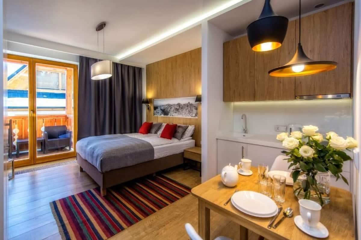 A modern hotel room with a bed, a dining table with breakfast items, a kitchen area, and a view through a glass door.