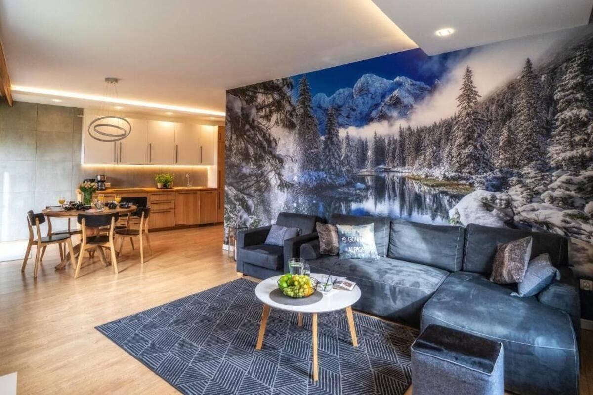 A modern living room with a large painting of a snowy mountain landscape, a dark grey sofa, and a round coffee table with fruits.