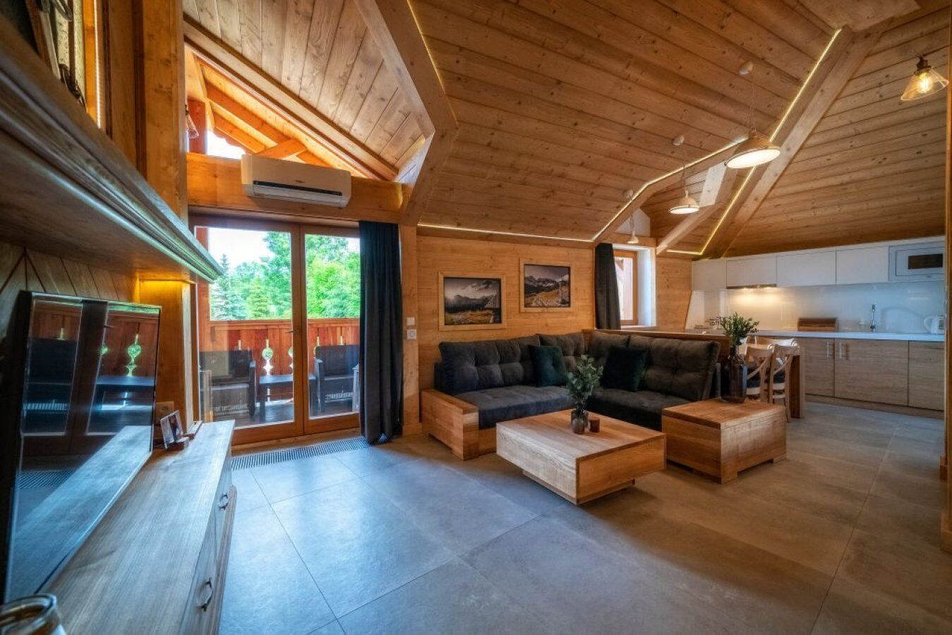 A cozy cabin interior with a wooden ceiling, a sofa, a coffee table, and a kitchen area.